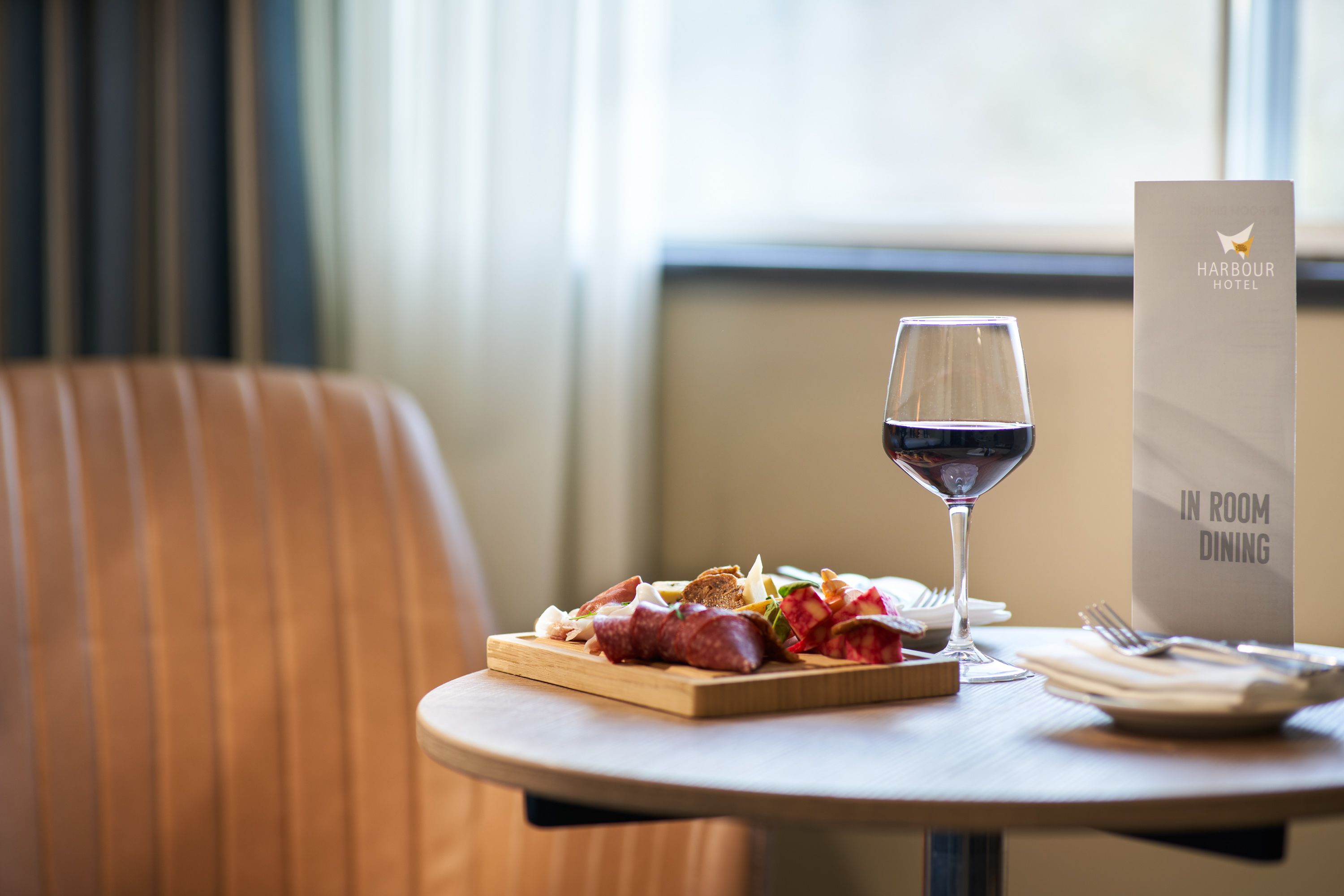 A dining table with a plate of food, a glass of red wine, and a sign indicating in-room dining at Harbour Hotel.