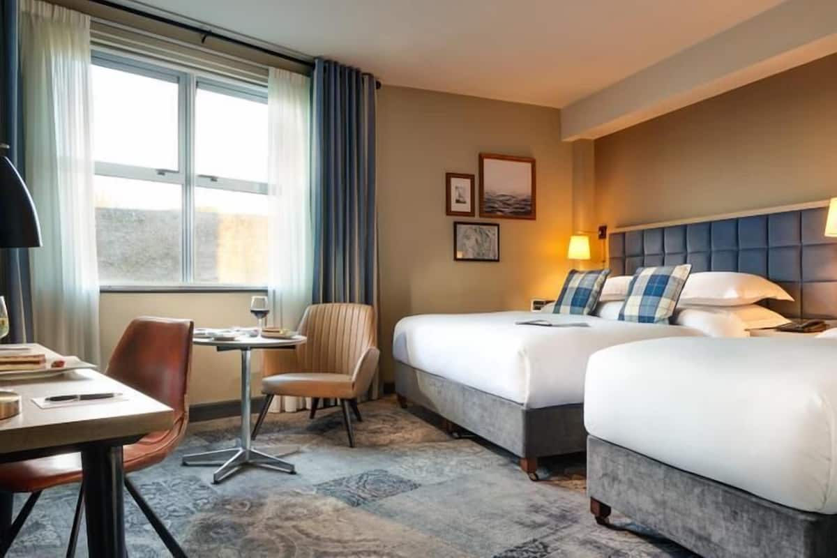 A hotel room with two beds, a desk with a chair, a small table, and a window with curtains.