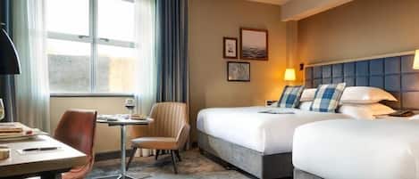 Habitación de hotel con dos camas, un escritorio con silla, una mesita y un ventanal con cortinas.