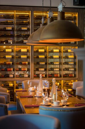 Una bodega con estantes llenos de botellas de vino, una mesa de comedor dispuesta con copas y cubiertos, y una lámpara colgante.