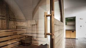 A modern sauna with wooden walls and a glass door, a wooden bench, and a small shelf with towels. - Engadiner Boutique-Hotel GuardaVal (Scuol)