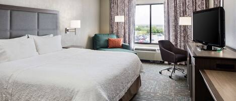 A hotel room with a large bed, a desk with a computer, a chair, and a window with curtains.