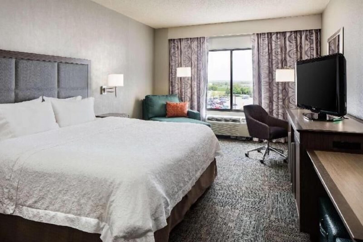 A hotel room with a large bed, a desk with a computer, a chair, and a window with curtains.