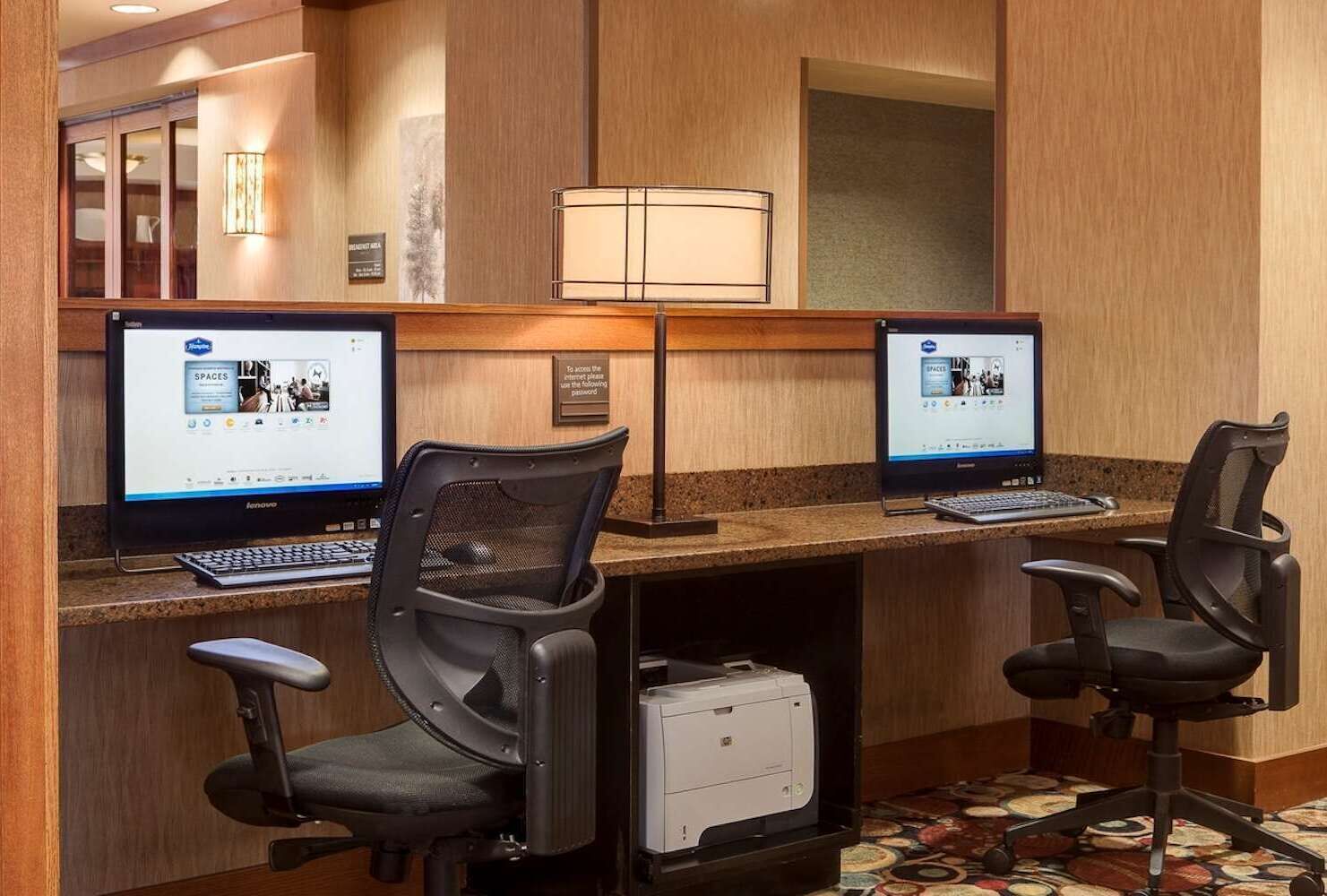 A hotel workspace with two Lenovo computers, two black office chairs, a printer, and a lamp.