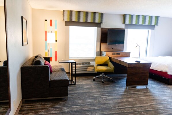 A hotel room with a sofa, a desk, a chair, a bed, a TV, and a window with curtains.