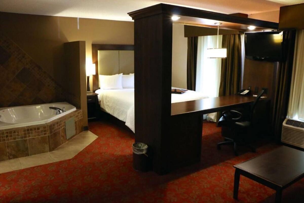 A hotel room with a large bed, a separate bathroom with a bathtub, a desk, a chair, and a flat-screen TV mounted on the wall.