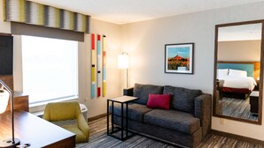 A hotel room with a grey sofa, a small table, a lamp, a mirror, and a picture on the wall.