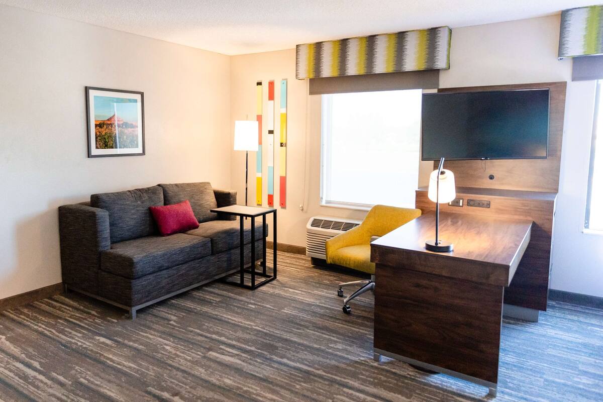 A modern hotel room with a gray sofa, a yellow chair, a wooden desk, a flat-screen TV, and a large window with a view.