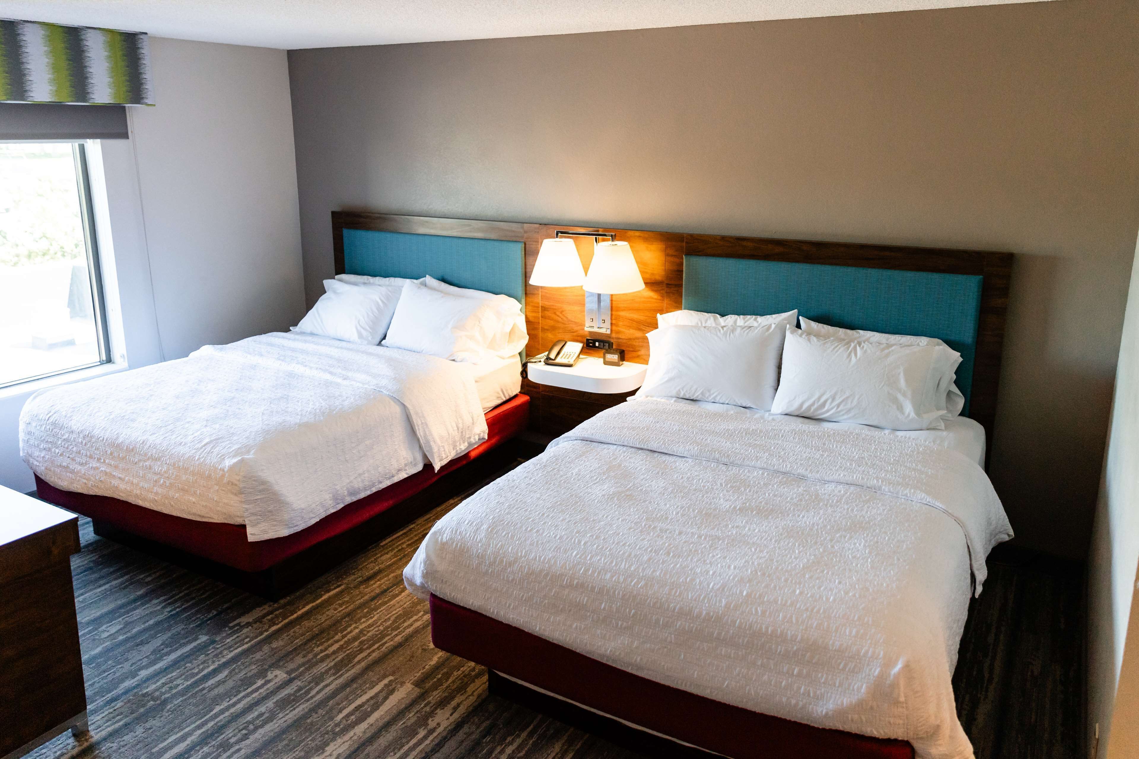 A hotel room with two beds, a nightstand with lamps, and a window providing natural light.