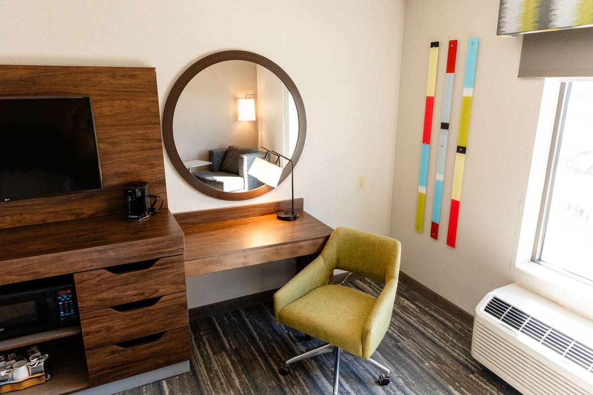 A hotel room with a wooden desk, a television, a round mirror, a green chair, a wall-mounted air conditioner, and colorful vertical blinds.