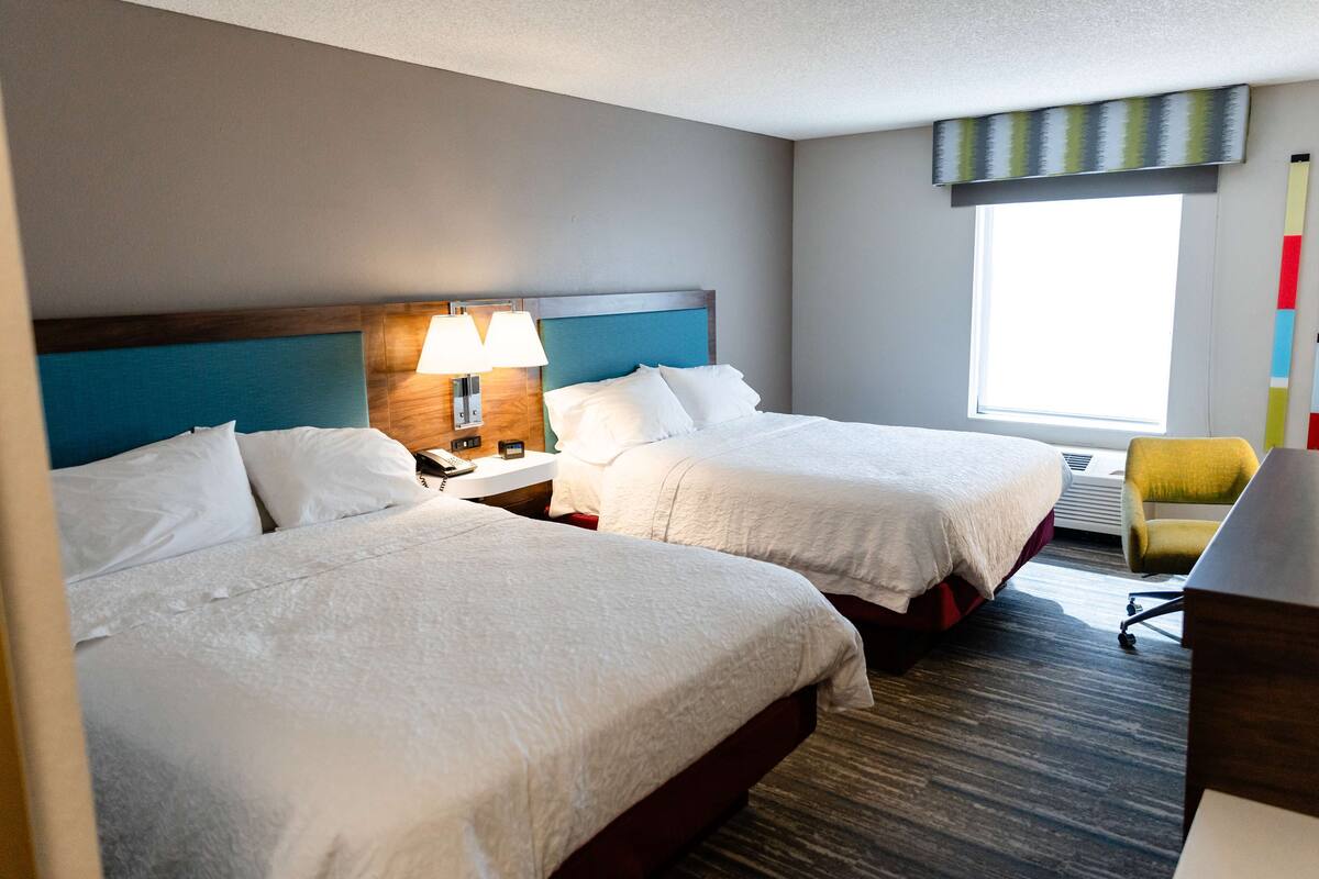 A hotel room with two beds, a desk, a chair, and a window with a view.