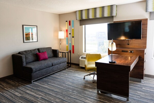A hotel room with a grey sofa, a desk, a TV, a lamp, and a window with a city view.