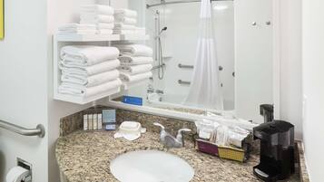 A bathroom with a granite countertop, a sink, a mirror, a towel rack, a shower, and a coffee maker.
