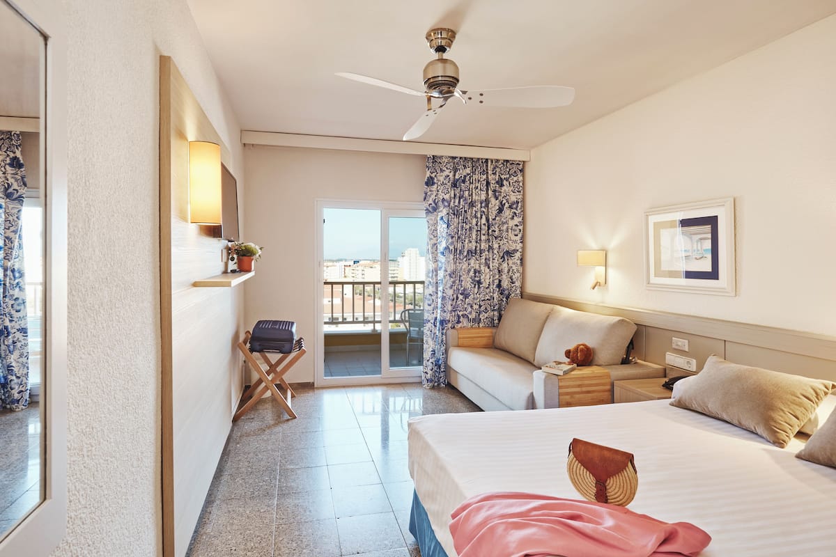 A hotel room with a bed, a sofa, a ceiling fan, a nightstand, a lamp, a window with curtains, a view of the city, and a door with a mirror.