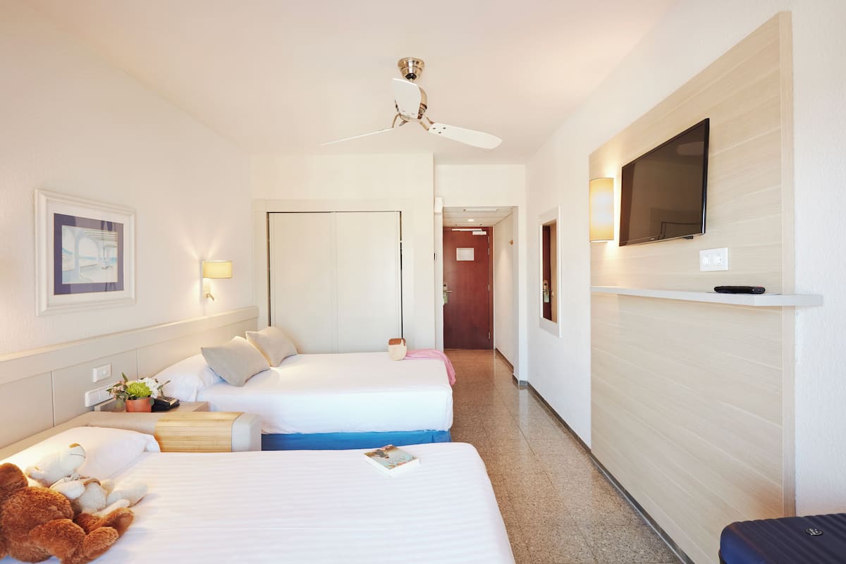 A hotel room with two beds, a ceiling fan, a TV mounted on the wall, and a small table with a lamp.