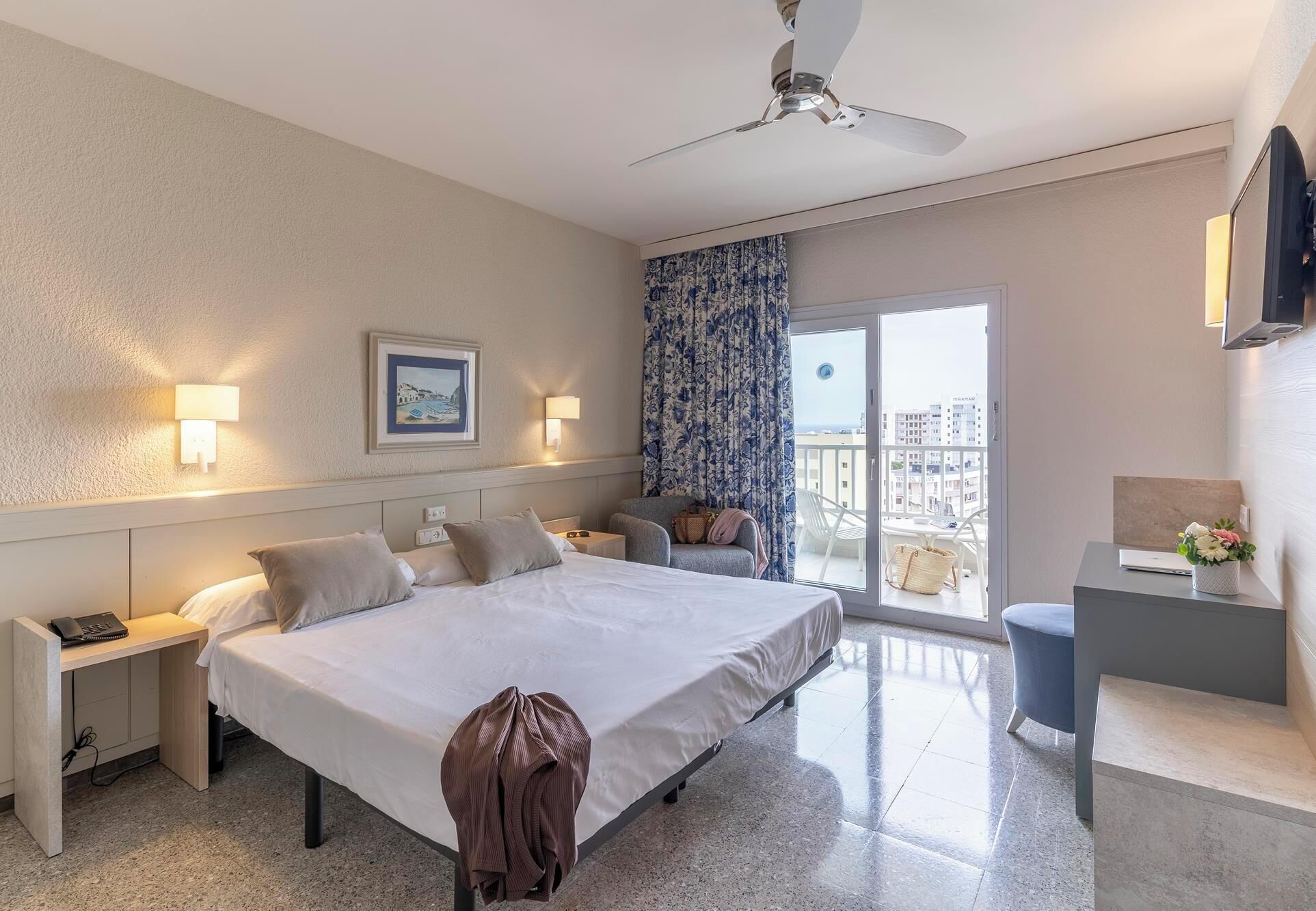 A hotel room with a bed, bedside tables, a desk, a chair, a ceiling fan, a TV, and a balcony with a view.
