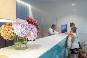 A reception desk with a vase of flowers, a digital screen, and a counter. - Prestige Goya Park (Roses)