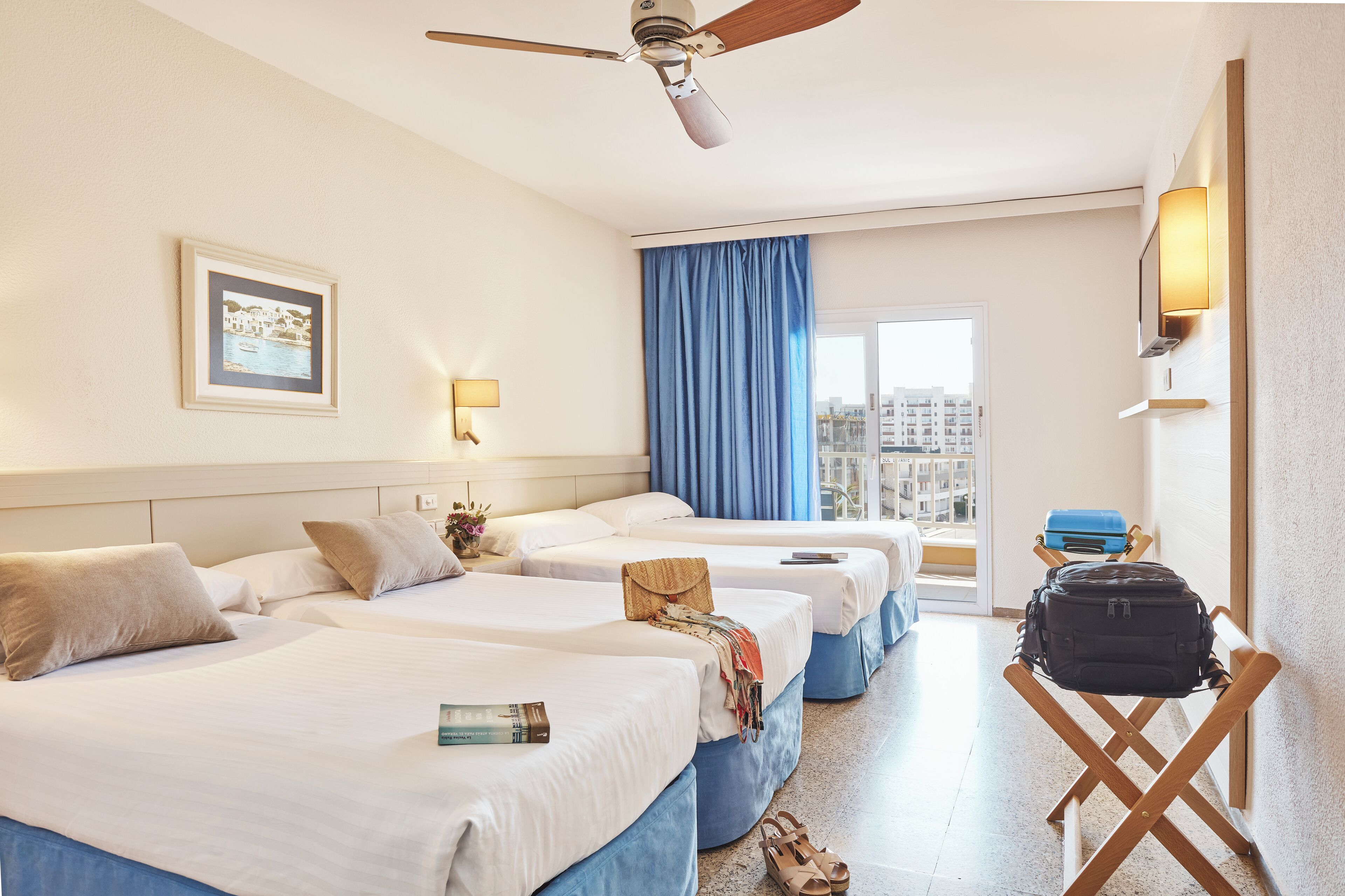 Une chambre d’hôtel avec trois lits, un ventilateur de plafond, une grande fenêtre avec des rideaux bleus et une vue sur les bâtiments environnants.