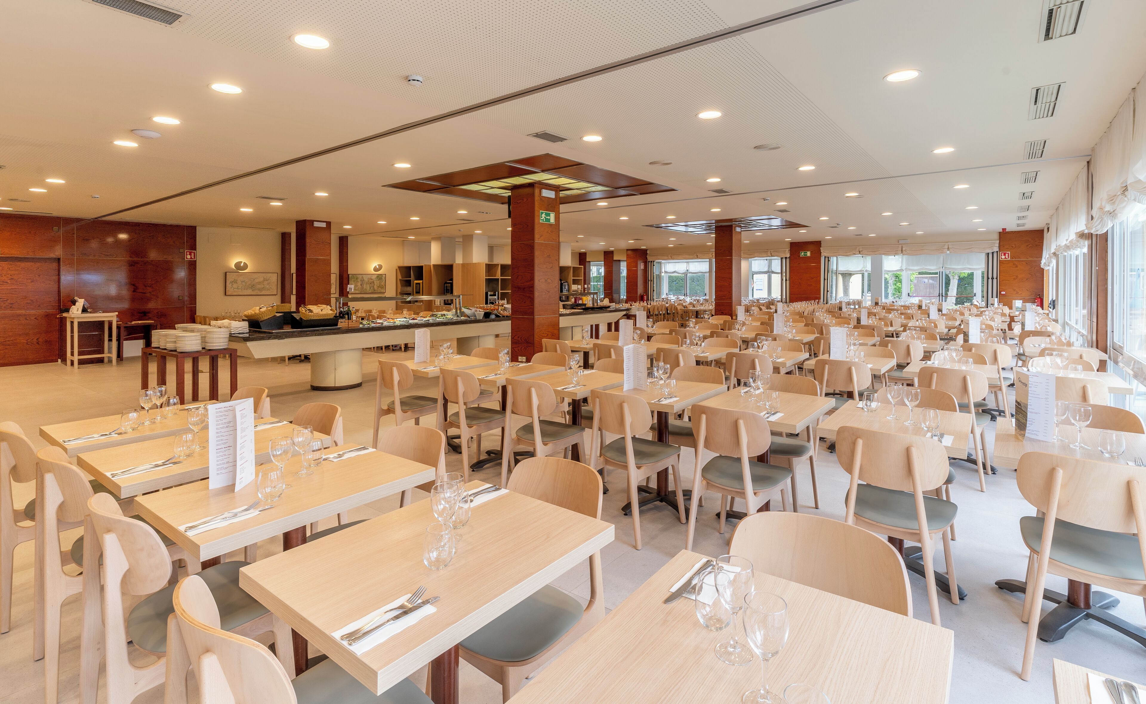 A spacious dining area with multiple tables and chairs, a buffet counter, and large windows allowing natural light.