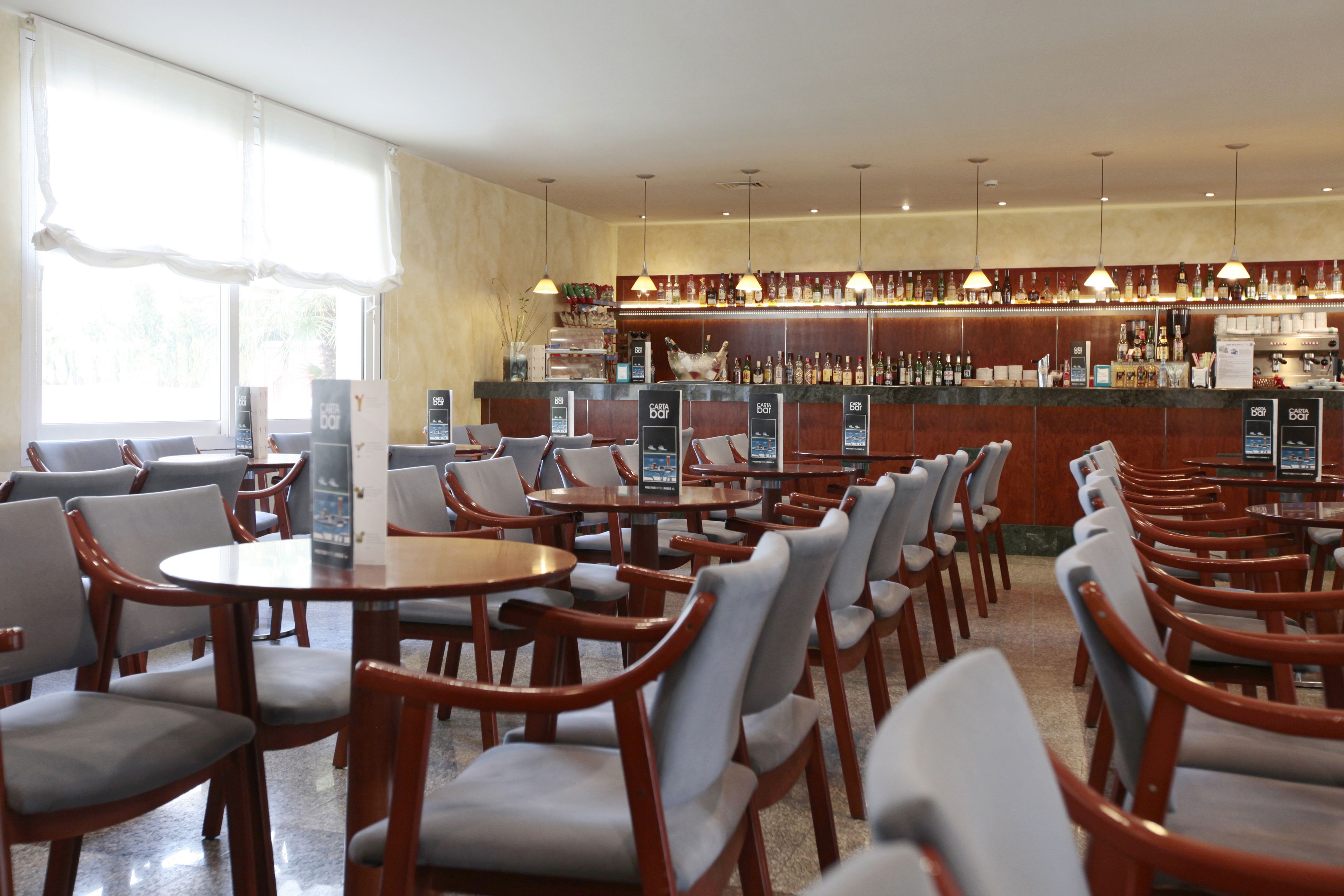 Zona de bar con varias mesas y sillas, una barra con diferentes botellas y un amplio ventanal que proporciona luz natural.