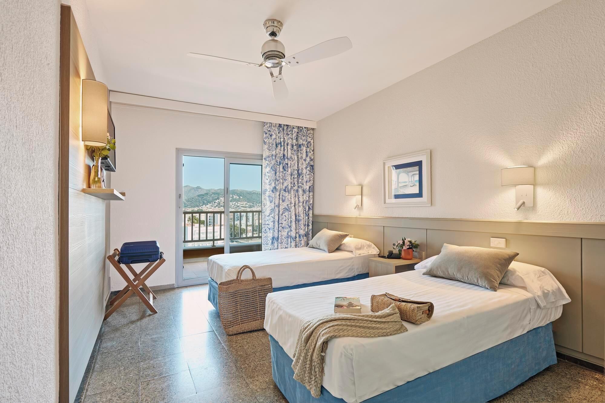 A hotel room with two beds, a ceiling fan, a balcony with a view, and a wall-mounted lamp.