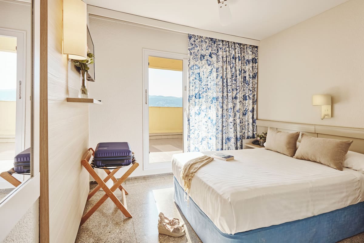 A hotel room with a bed, a suitcase on a chair, a nightstand, and a view through a door.