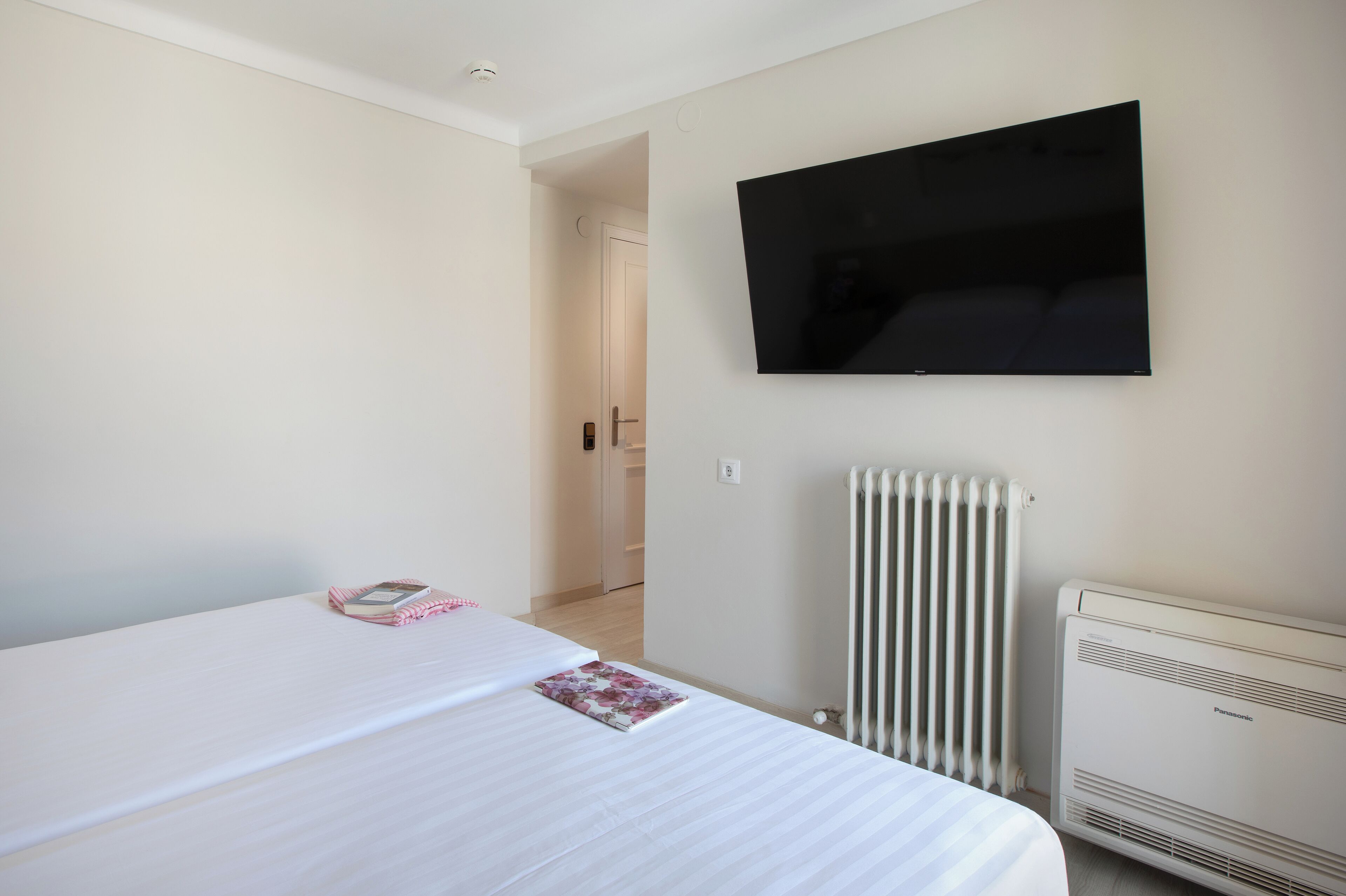 Un dormitorio con cama, televisor de pantalla plana montado en la pared, radiador y puerta que da a otra habitación.