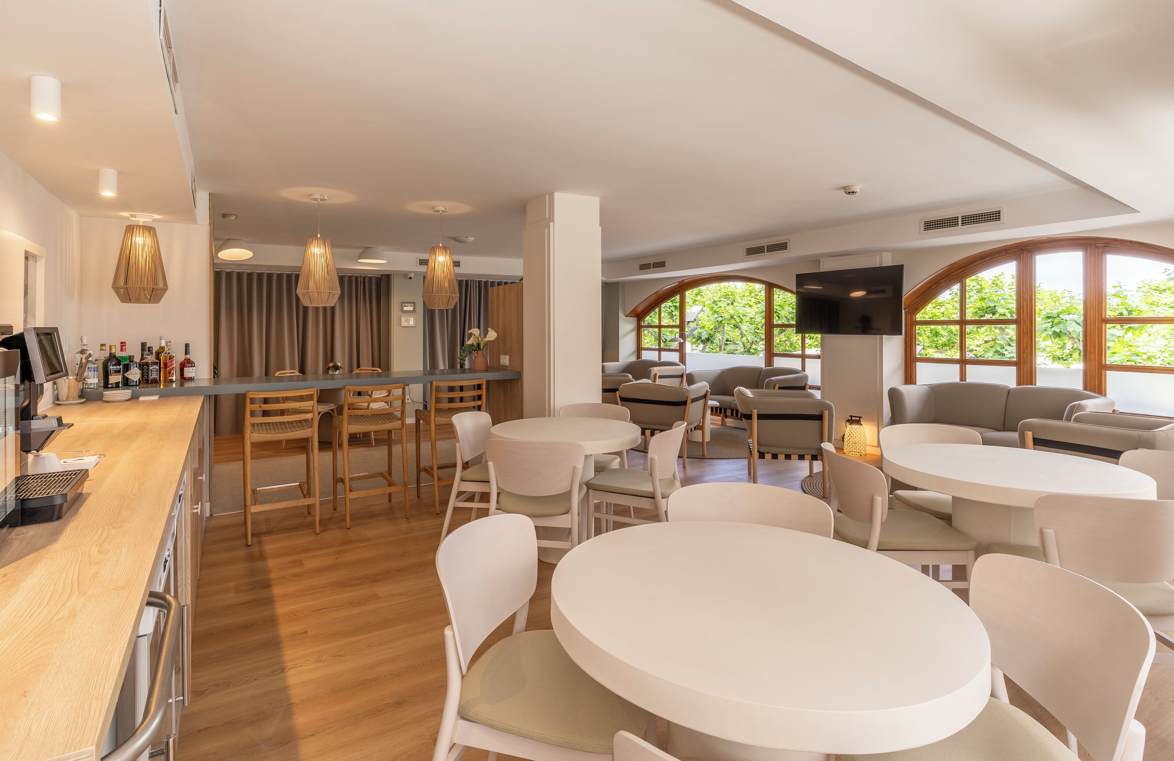 Un hall d’hôtel moderne, avec des tables et des chaises rondes, un bar où sont exposées des bouteilles, et de grandes fenêtres donnant sur un paysage verdoyant.