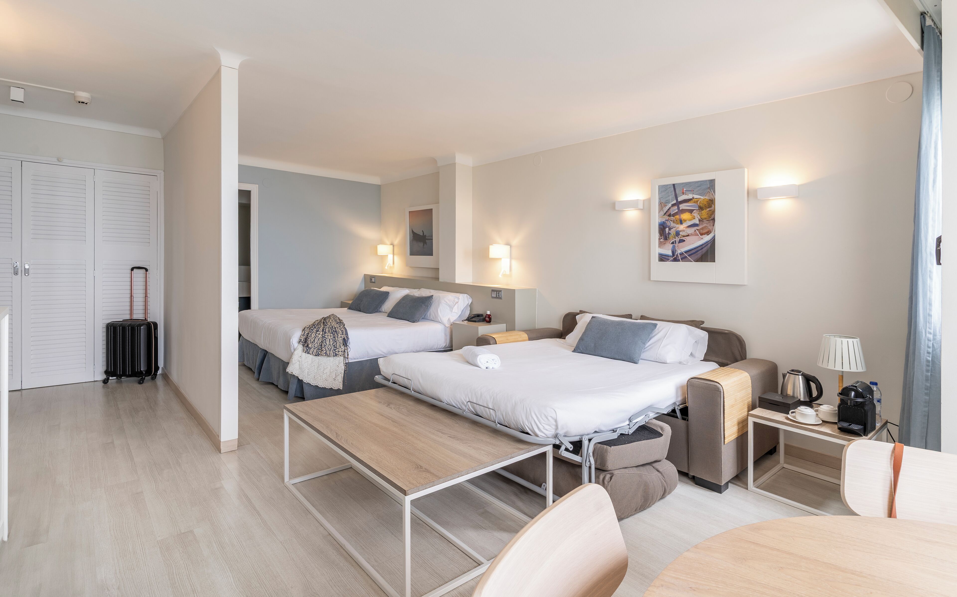 A hotel room with two beds, a desk, a chair, a suitcase, and a coffee machine.