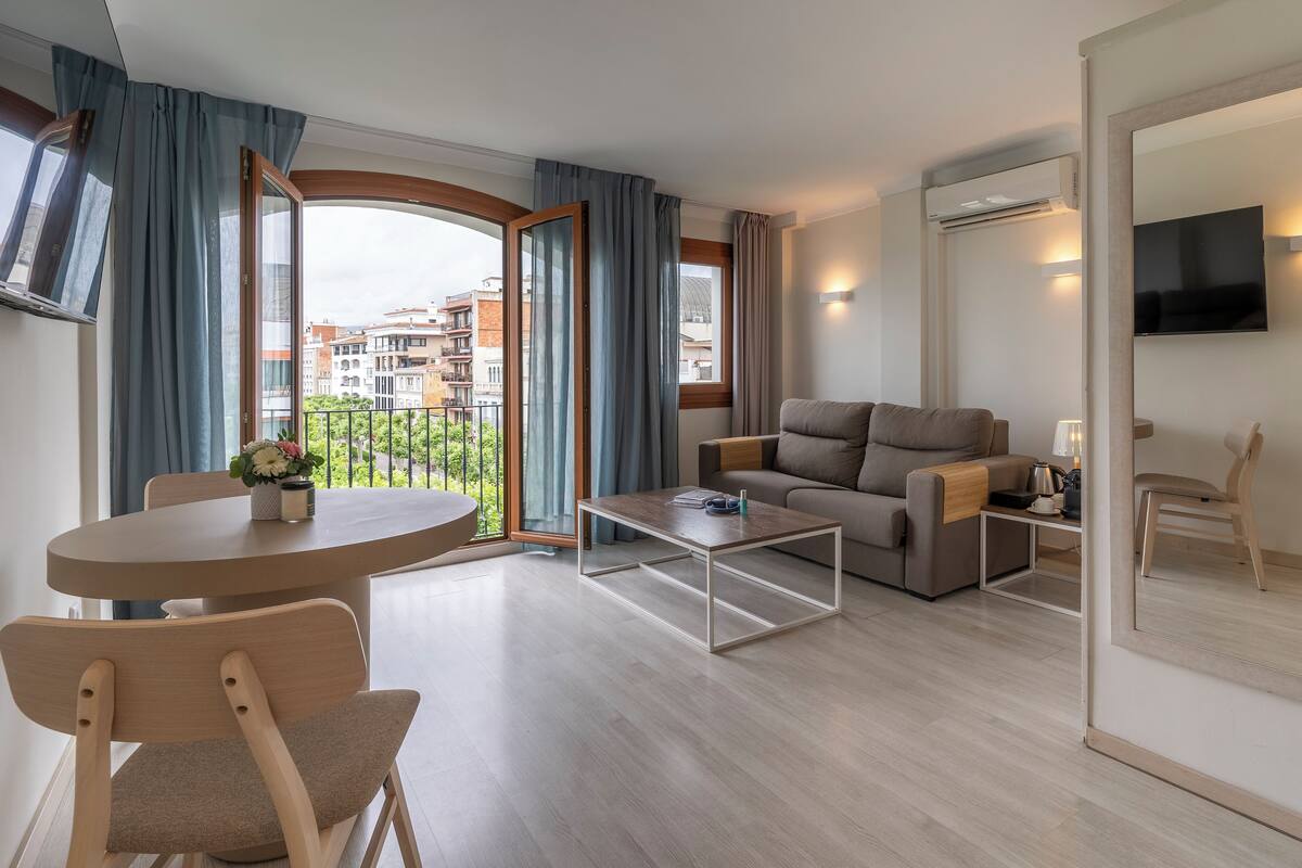Un salon moderne avec un canapé, une table basse et des fauteuils, un balcon avec des rideaux et un téléviseur fixé au mur.
