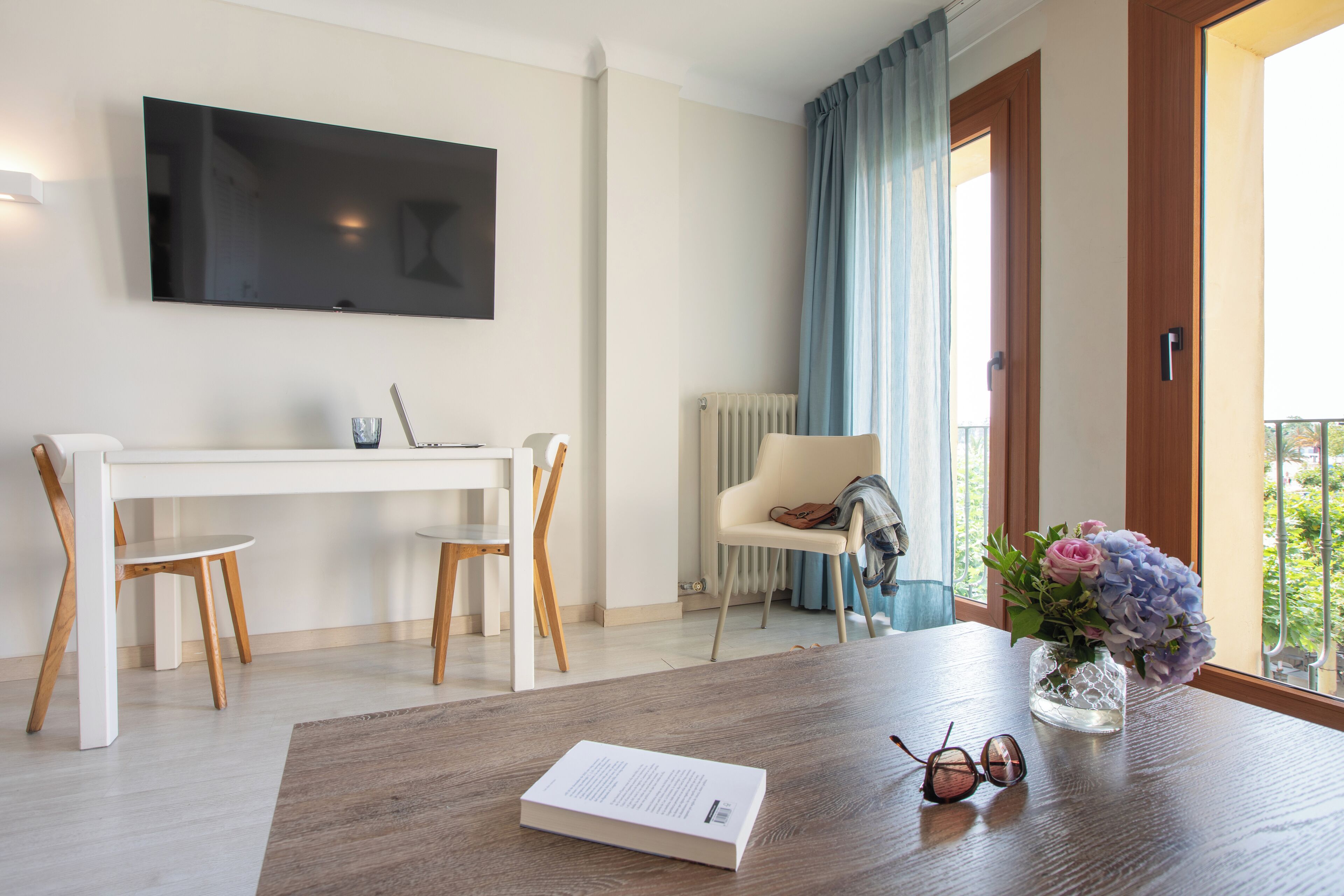 A modern room with a wooden dining table, a white desk, two wooden chairs, a TV mounted on the wall, a radiator, a window with blue curtains, and a vase with flowers on the table.