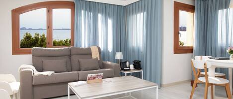 A modern living room with a sofa, coffee table, and dining area, featuring large windows with a view of the water.