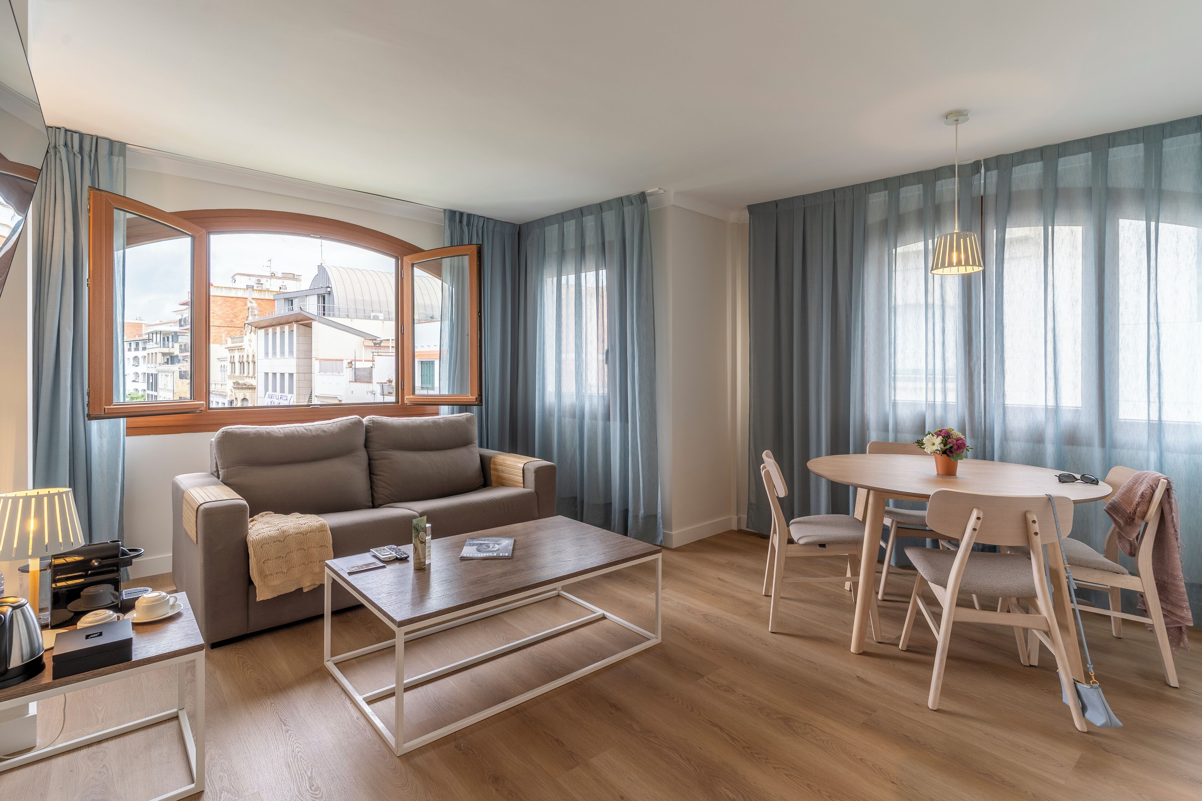 A modern living room with a sofa, coffee table, and dining table. A large window offers a view of buildings outside.