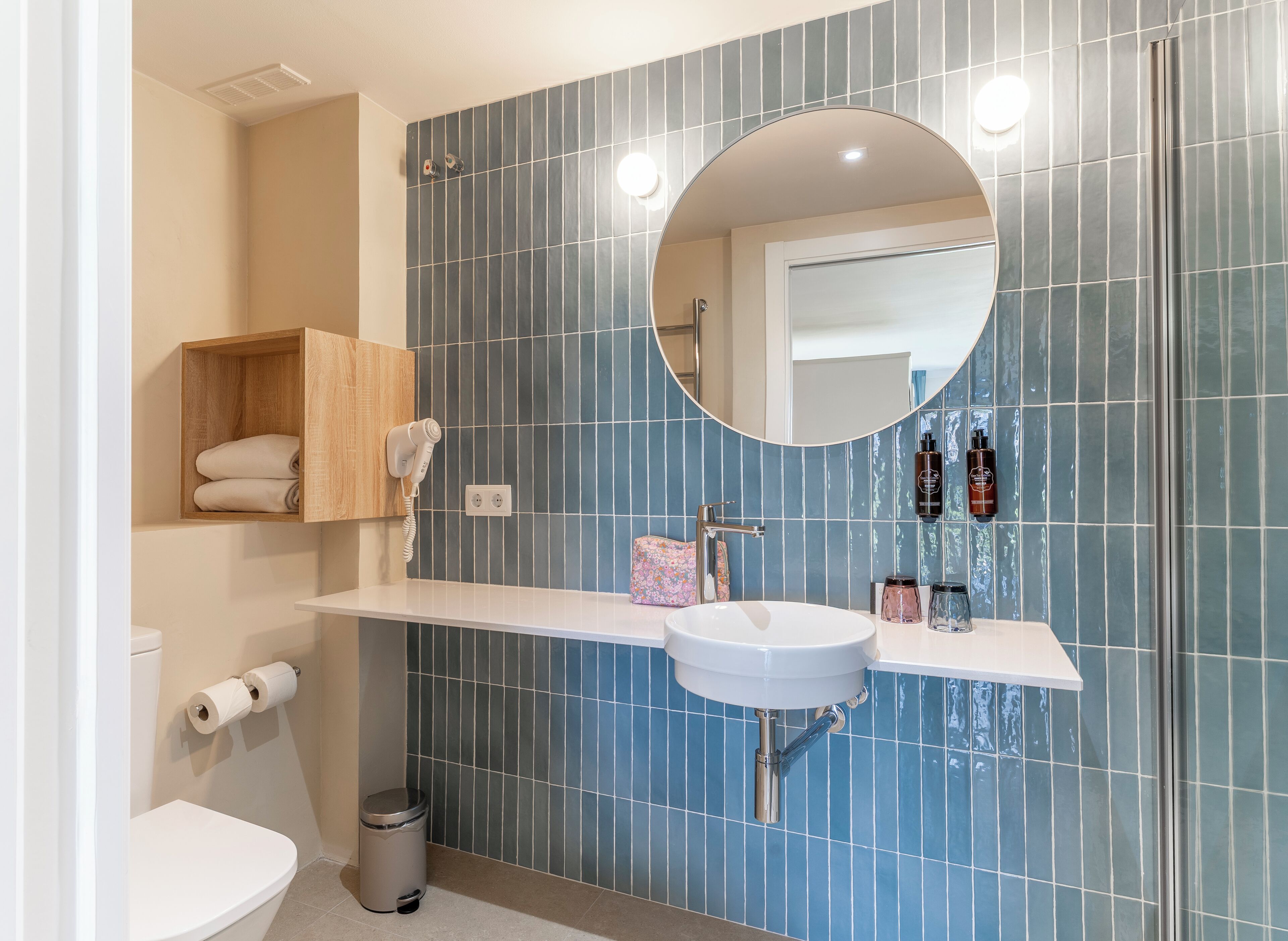 Ein modernes Badezimmer mit einem weißen Waschbecken, blauen Fliesen, einem Holzbodenregal mit Handtüchern, einer Toilette und einem Papierkorb.