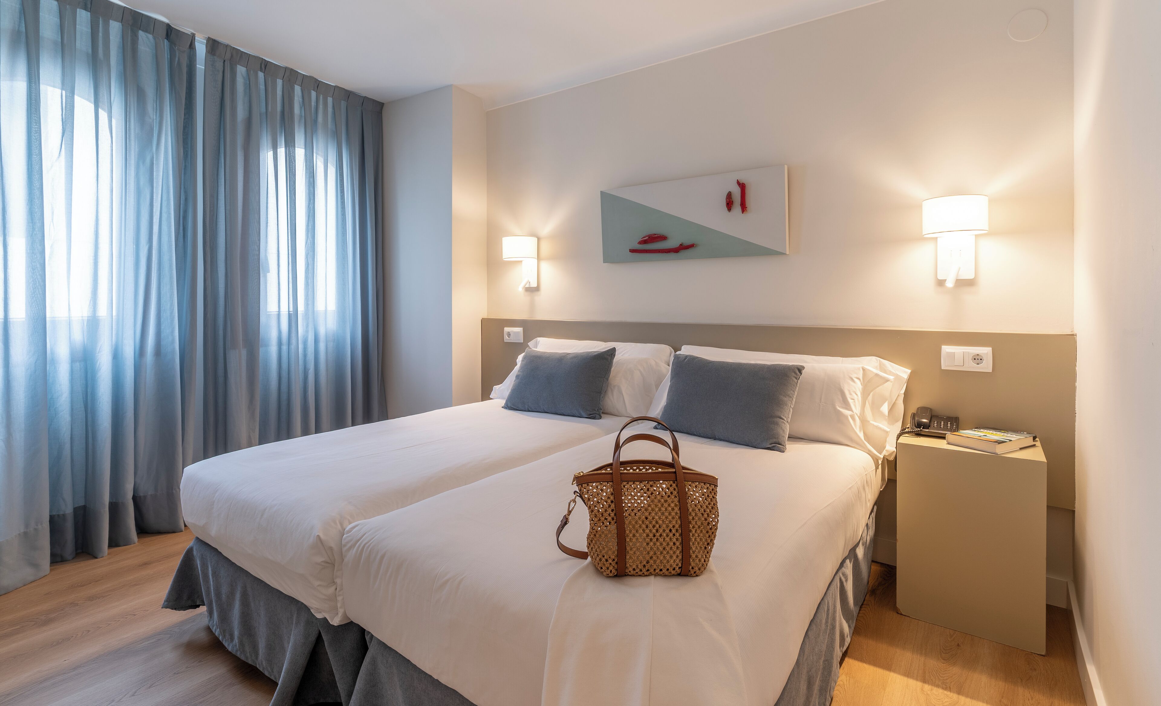 Habitación de hotel con una cama grande, dos lámparas de noche, una mesita de noche, un bolso tejido y un cuadro en la pared.