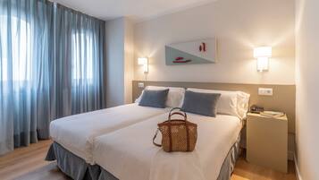 Habitación de hotel con una cama grande, dos lámparas de noche, una mesita de noche, un bolso tejido y un cuadro en la pared.