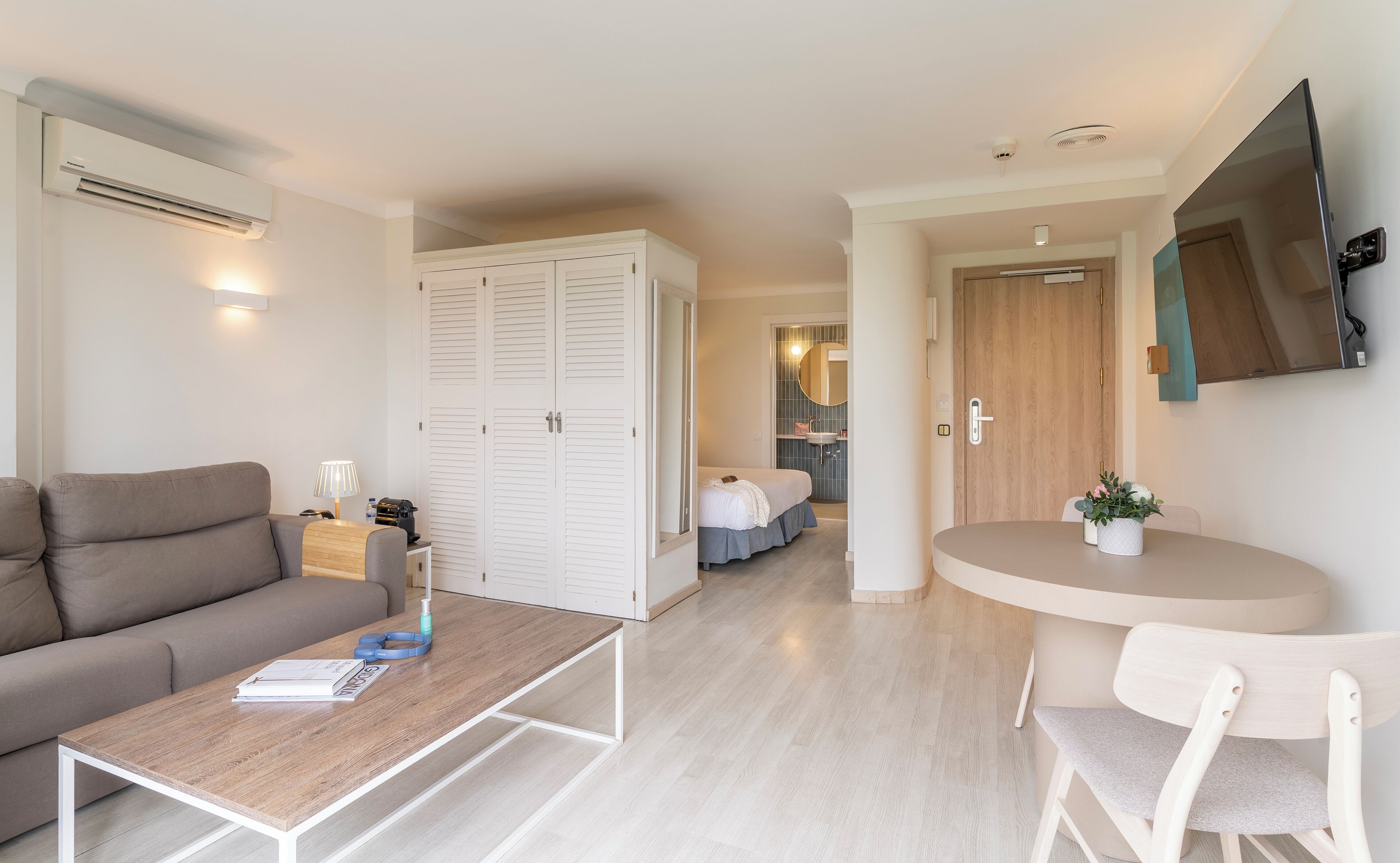 A modern hotel room with a sofa, a small table, a bed, a bathroom, and a TV mounted on the wall.