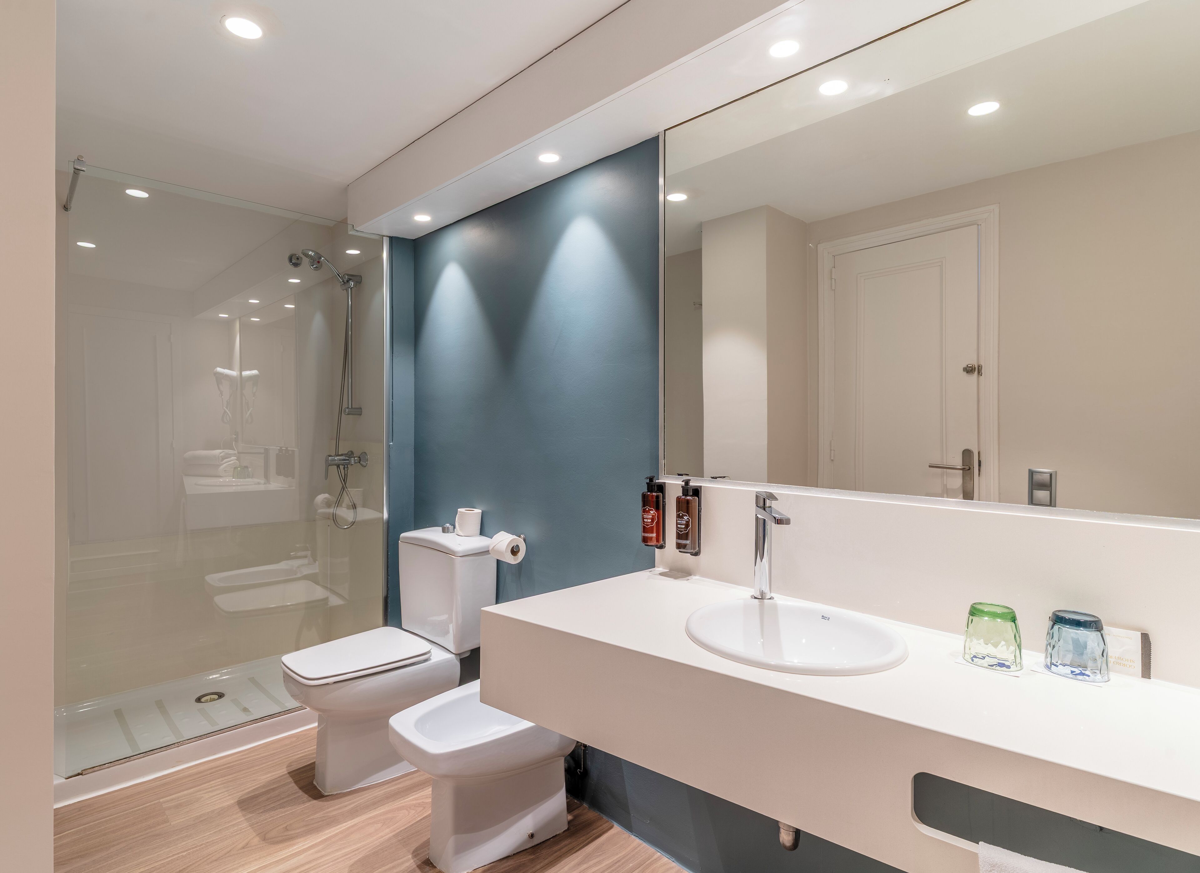 A modern bathroom with a glass-enclosed shower, a white toilet, a bidet, and a large mirror above a white sink with two soap dispensers.