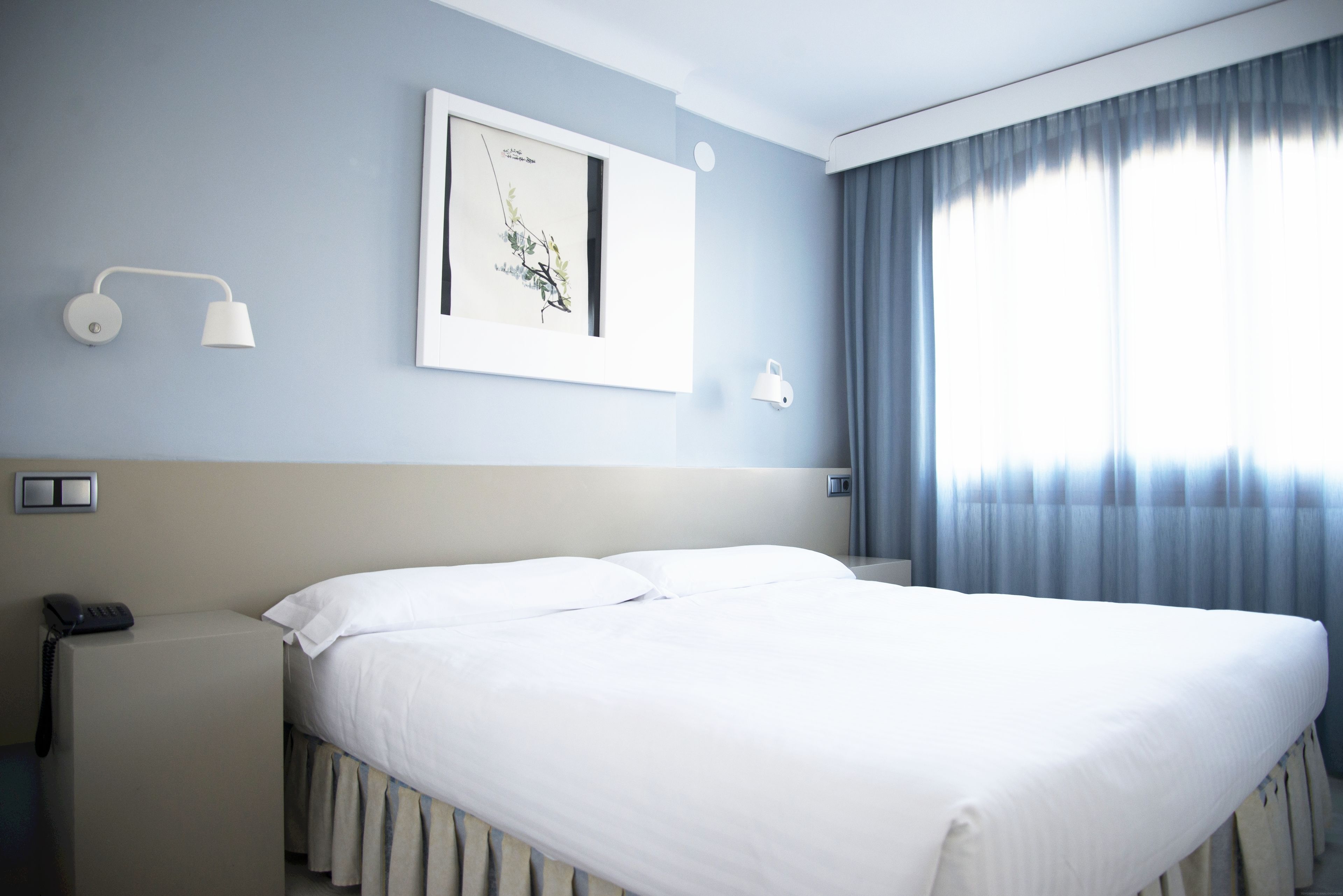 A hotel room with a bed, bedside table, lamp, and artwork on the wall.