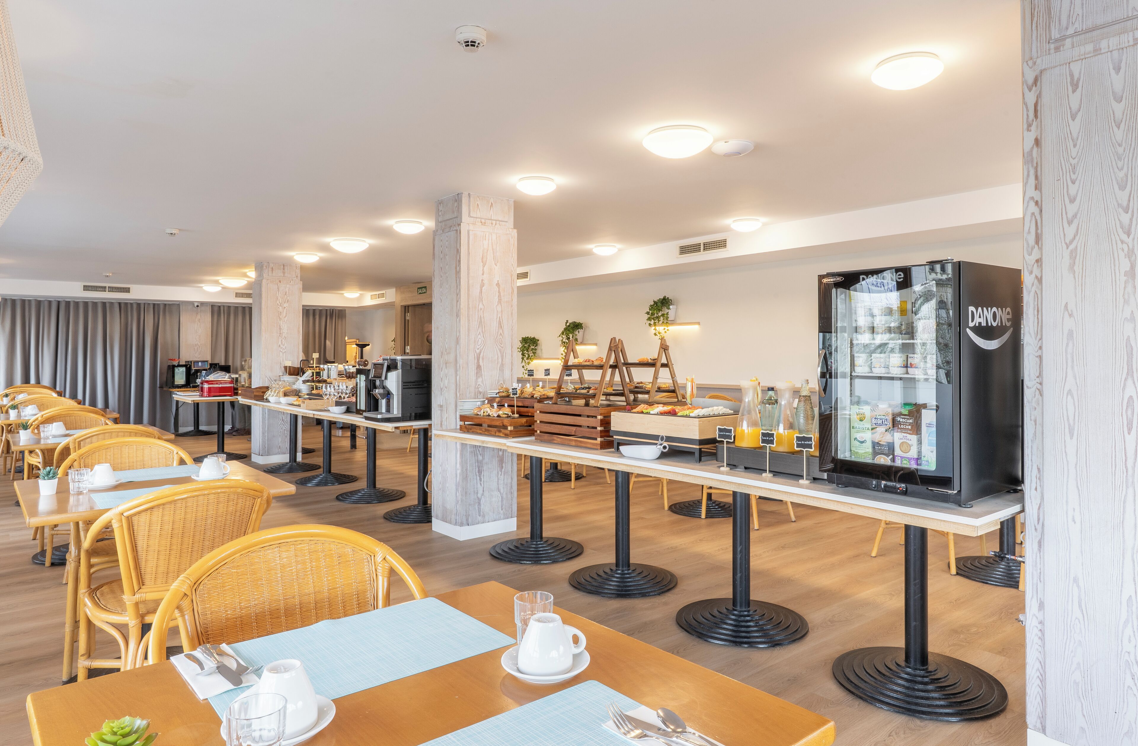 A modern hotel dining area with wooden tables, wicker chairs, and a central buffet station.