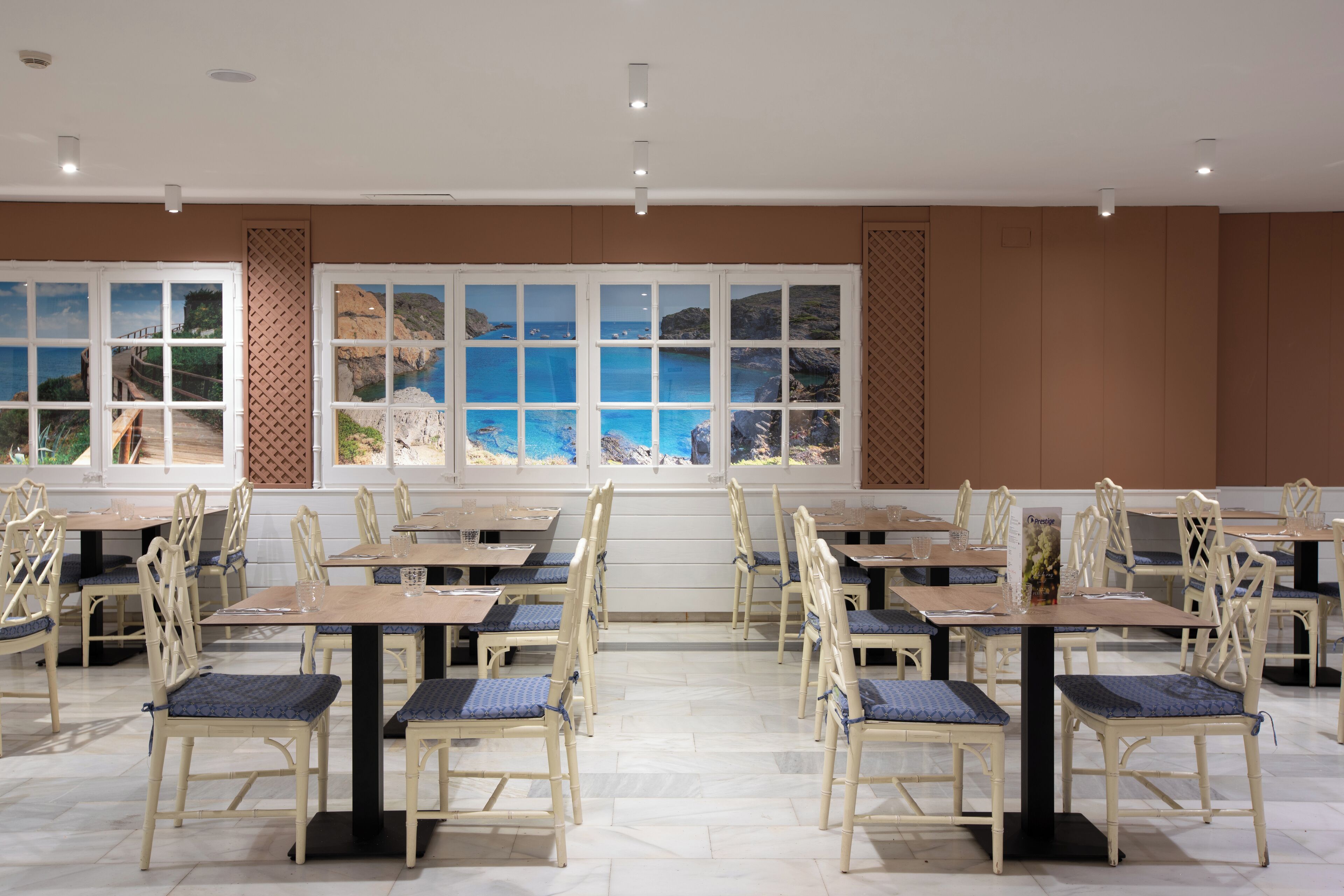 Un comedor con mesas y sillas, ventanas amplias con vistas al mar y un muro decorado.
