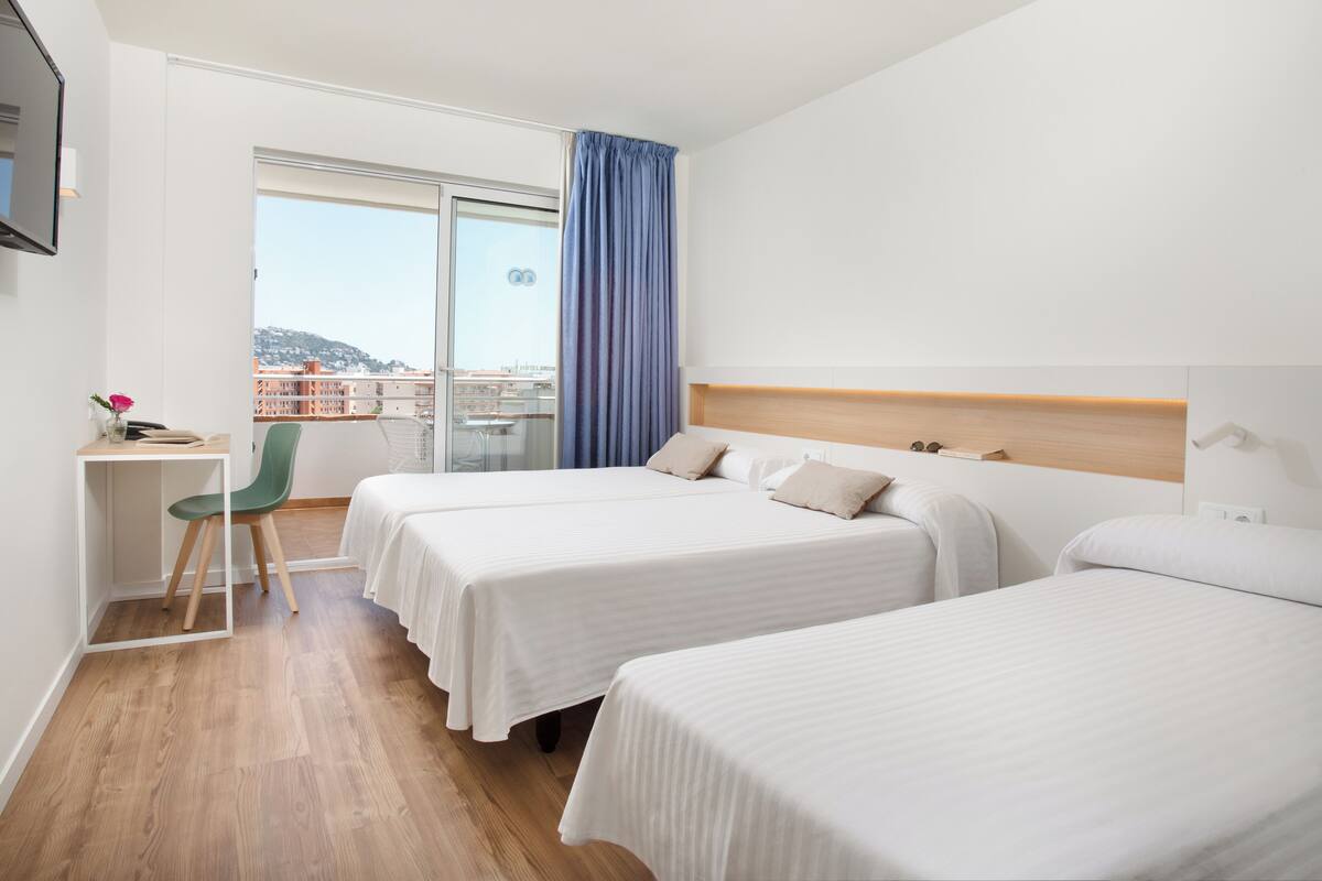 A hotel room with two beds, a desk, a chair, and a large window with a view of buildings and hills.