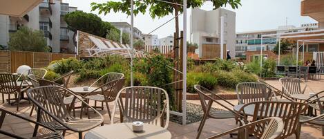 An outdoor patio area with multiple tables and chairs, surrounded by greenery and modern buildings in the background.