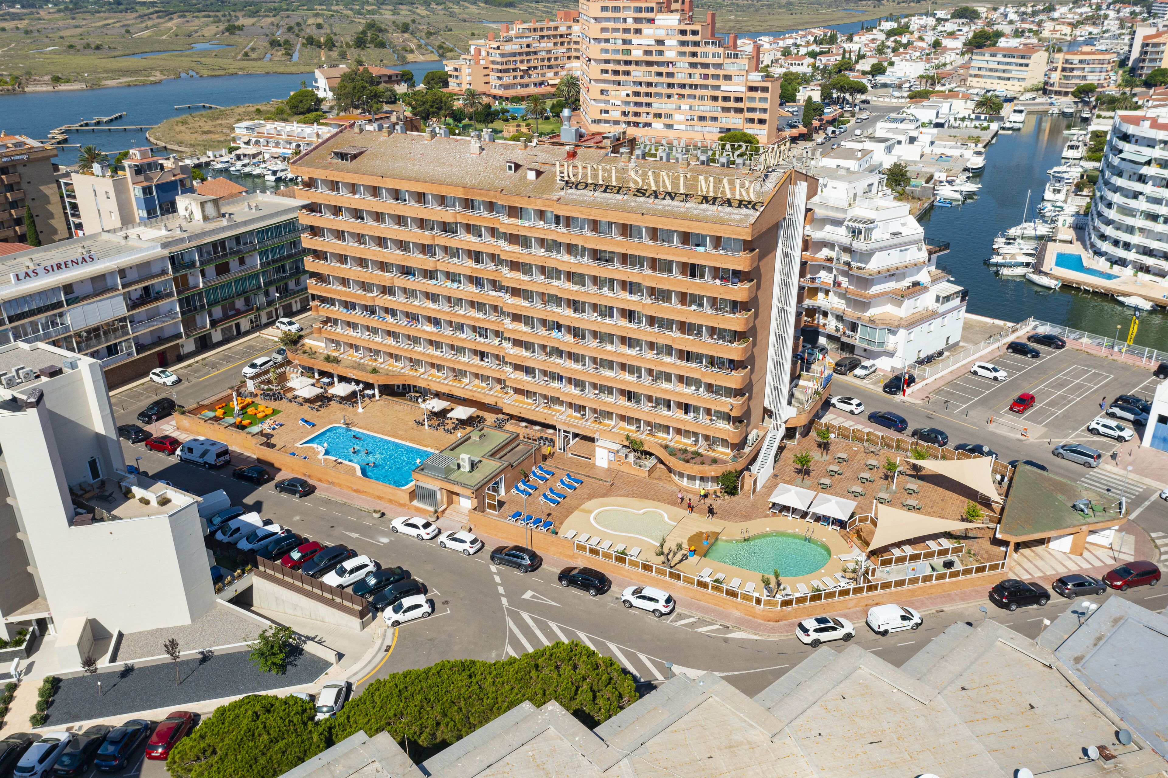 Un hotel de varios pisos con piscina en la azotea, un puerto deportivo con barcos y un estacionamiento.