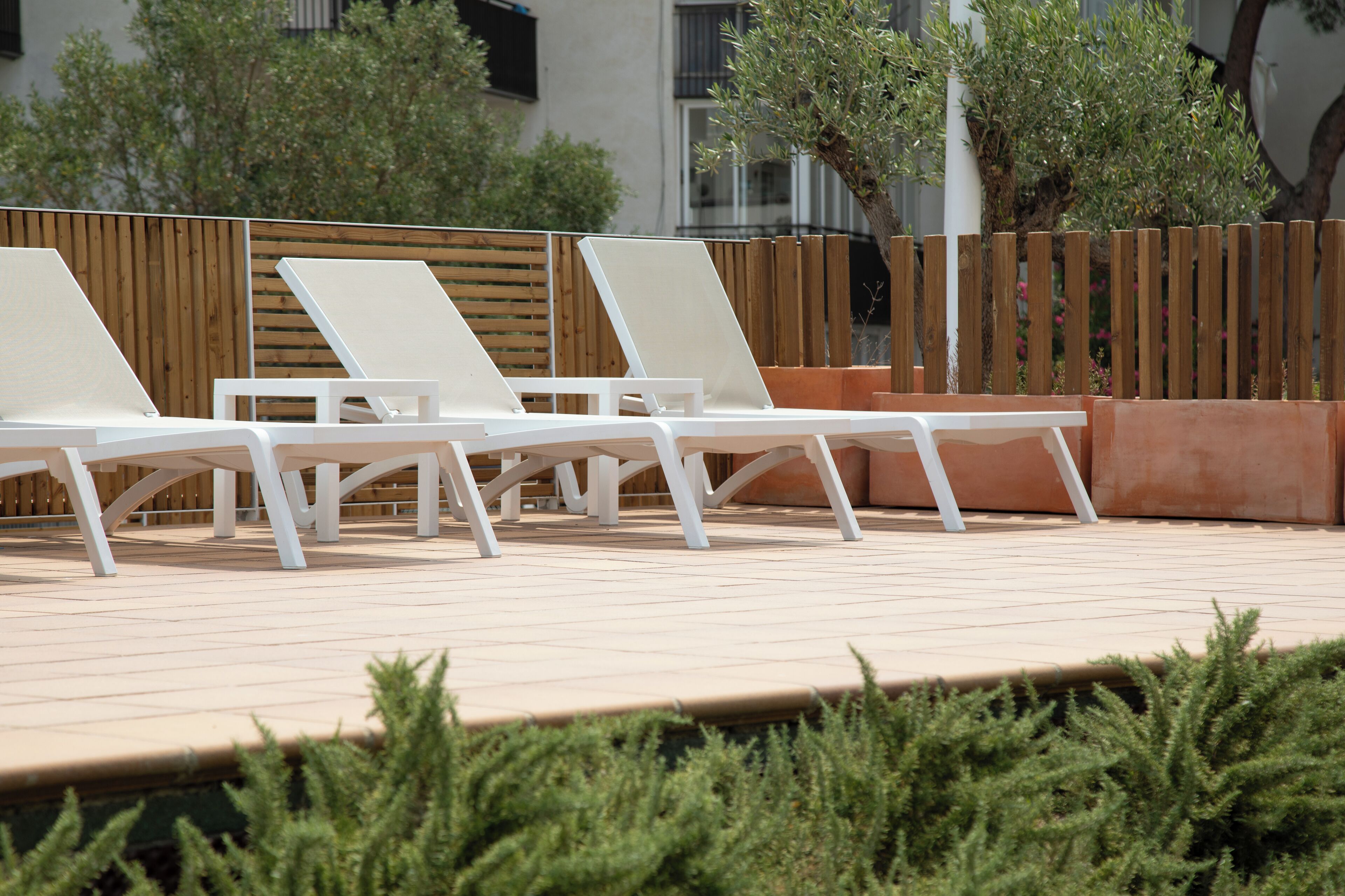 Una fila de sillas blancas para relajarse en una terraza con baldosas, una cerca de madera y follaje al fondo.