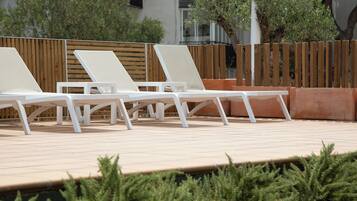 Una fila de sillas blancas para relajarse en una terraza con baldosas, una cerca de madera y follaje al fondo.