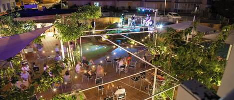 Eine Terrassenfläche im Freien mit Tischen und Stühlen, umgeben von viel Grün und einer Bühne für Auftritte.