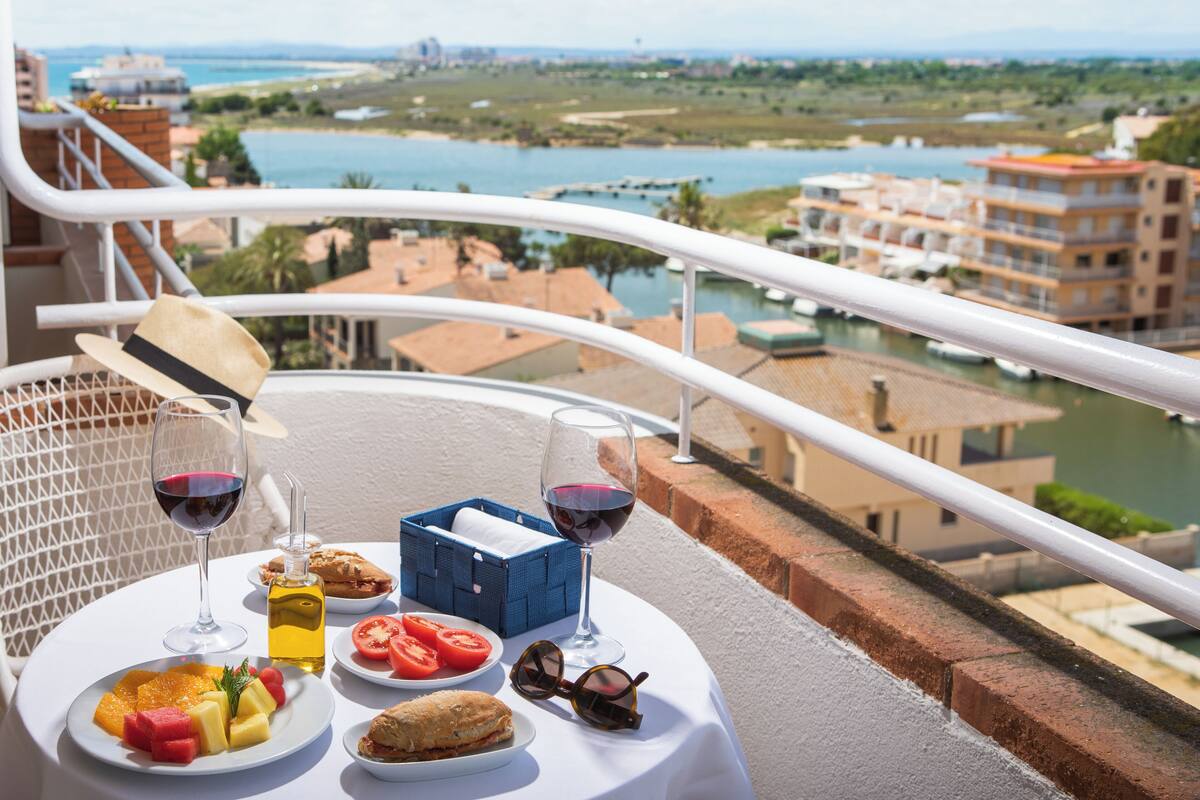 A balcony table with a view of the sea, a bottle of olive oil, a glass of red wine, a basket of bread, and a plate of fruit.