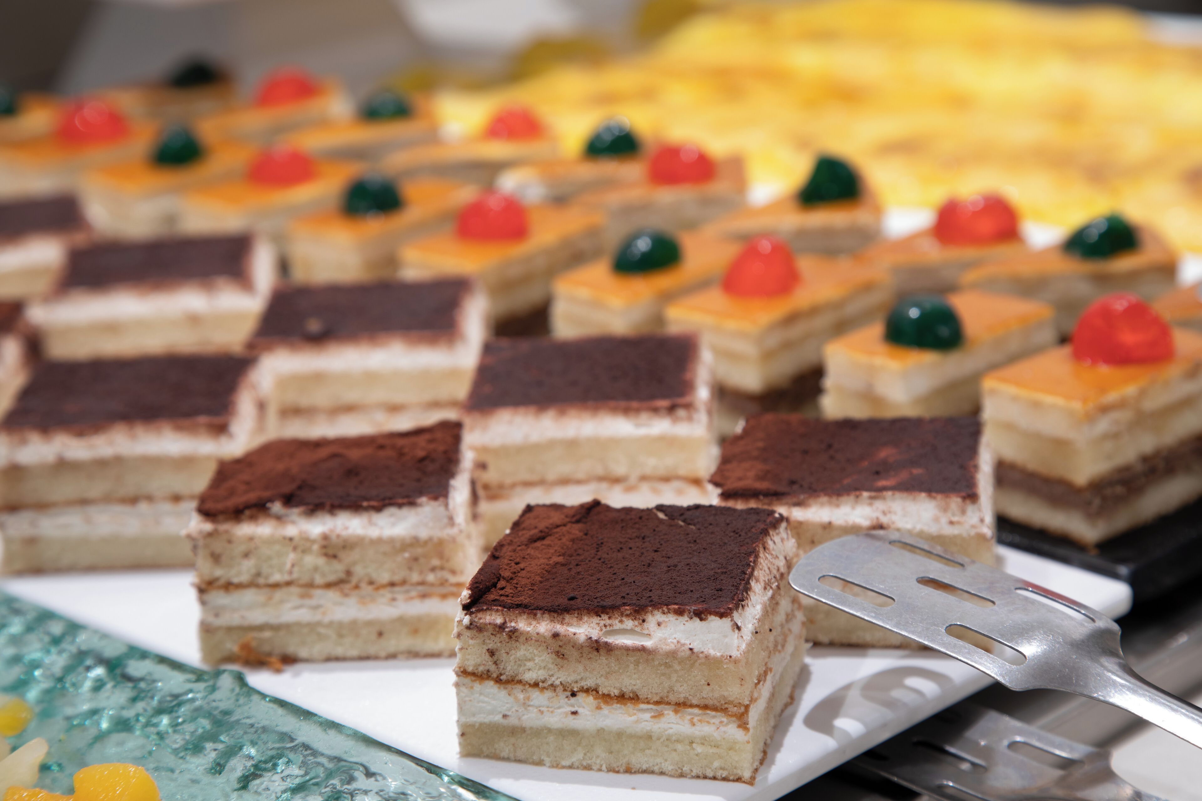 Un plat amb postres quadrades de diferents capes, algunes cobertes amb esferes de gelatina de colors, i una espàtula metàllica a un costat.