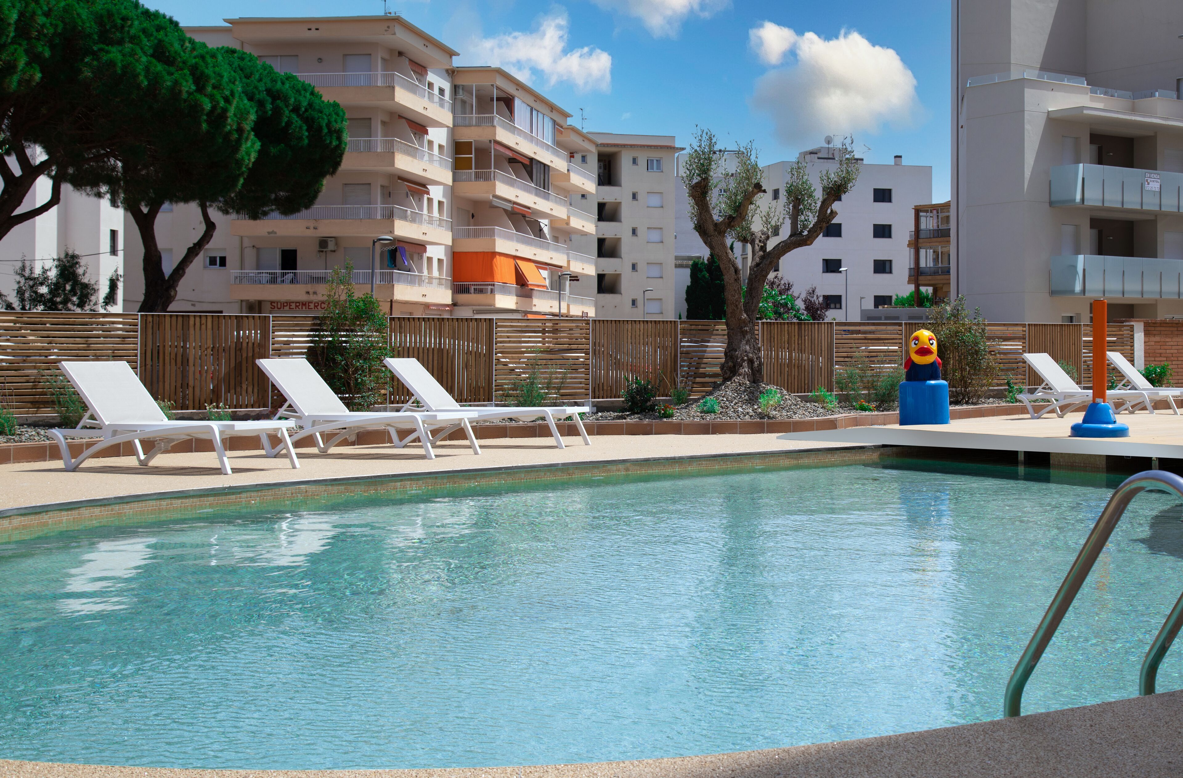 Une piscine avec des chaises longues, une clôture en bois et, au fond, des immeubles à plusieurs étages.